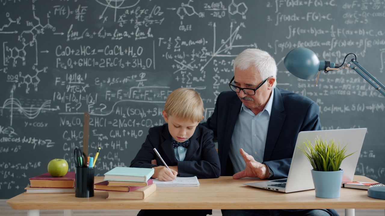 A senior tutor assists a young student with math homework at a desk in a classroom.