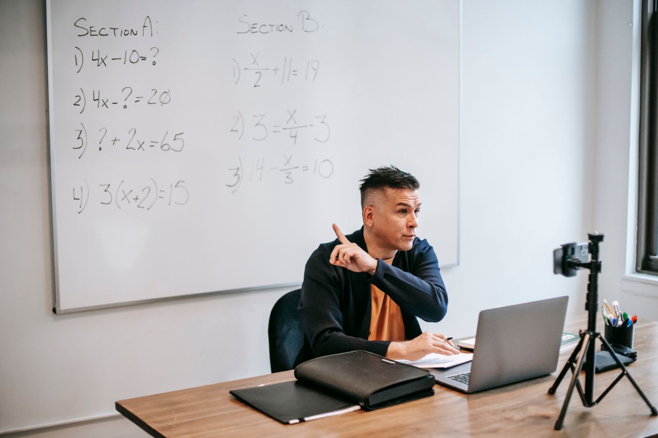 A male teacher explains math equations during an online class, using a laptop and whiteboard.